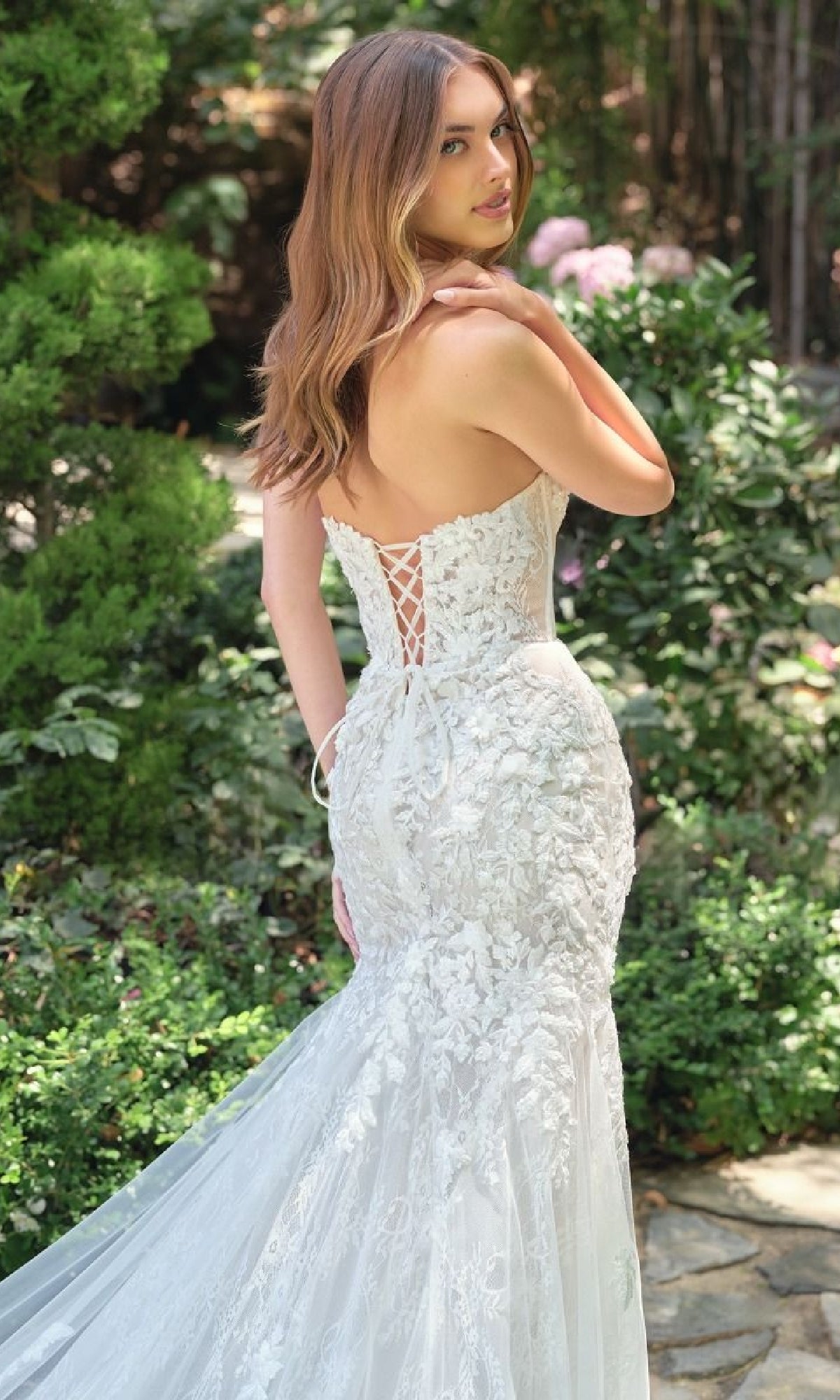 A woman in the Andrea & Leo WL088 strapless mermaid bridal dress with a lace-up back stands outdoors on a stone path, surrounded by greenery.